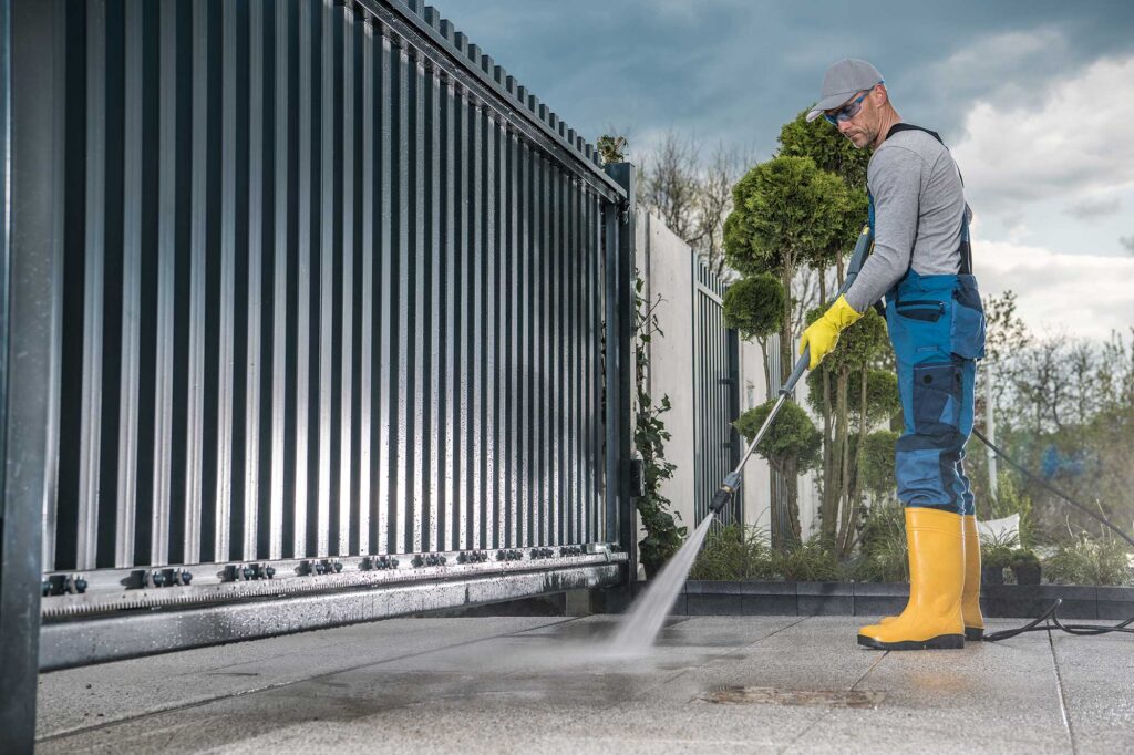 A worker in protective gear uses a pressure washer to clean a driveway next to a metal fence. The scene is set outdoors with greenery in the background.