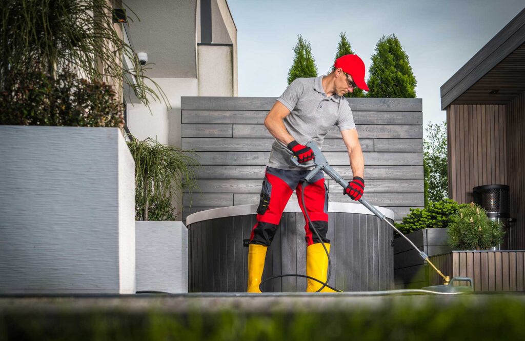 A person in a red cap and gloves uses a pressure washer to clean a patio area in a modern outdoor setting. The background features greenery and a stylish wooden wall.