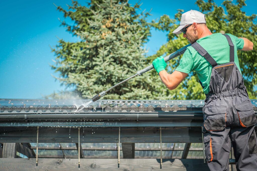 A worker cleans a roof using a pressure washer, surrounded by green trees and a clear blue sky. The scene emphasizes maintenance and outdoor work.