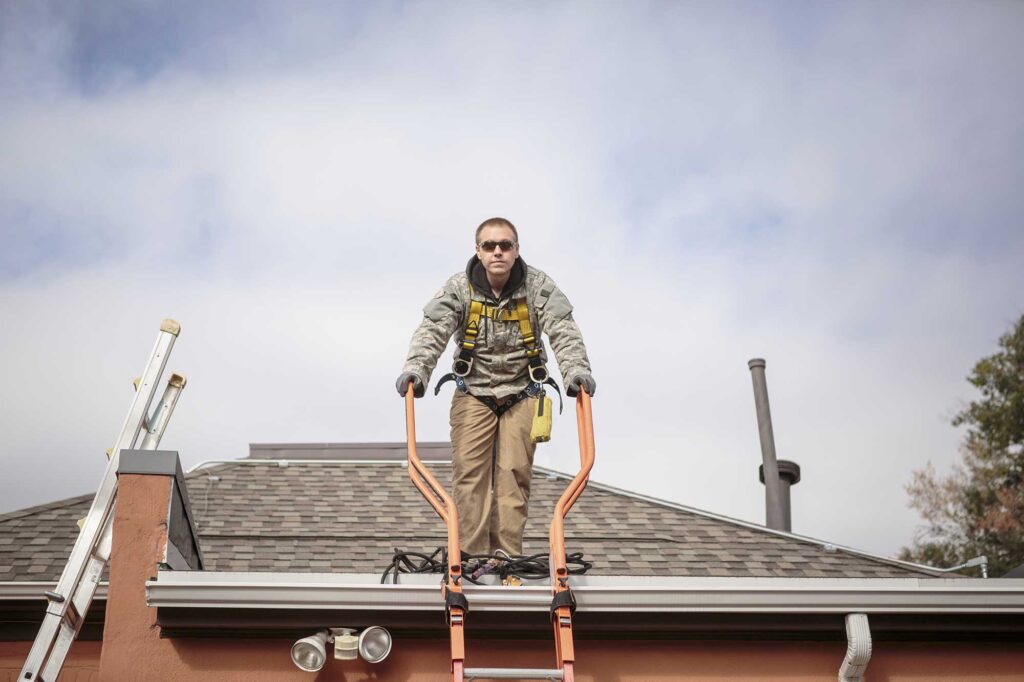 A worker balances on a roof using a specialized ladder, wearing safety gear and focused on the task. The sky is partly cloudy, indicating a typical day for outdoor maintenance work.
