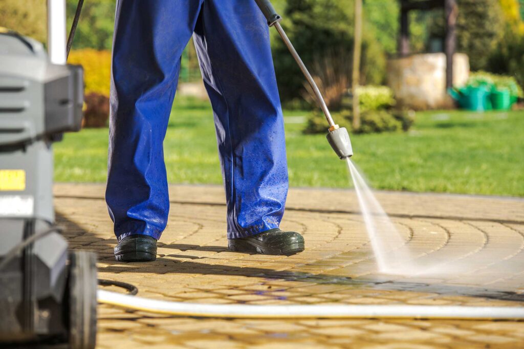 A person in blue work pants uses a pressure washer to clean a stone surface in a garden. The background features greenery and landscaping elements.